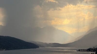 Dramatischer Himmel mit Sonnenstrahlen über einem See und Bergkette bei bewölktem Wetter