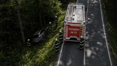 Feuerwehrleute stehen neben einem Einsatzfahrzeug an einer Straße, ein Auto liegt im Wald.