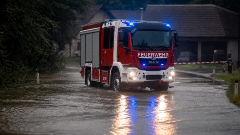 Rotes Feuerwehrauto mit Blaulicht fährt durch überflutete Straße bei Dämmerung