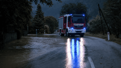 Rotes Feuerwehrfahrzeug mit Blaulicht fährt durch überflutete Landstraße bei Dämmerung