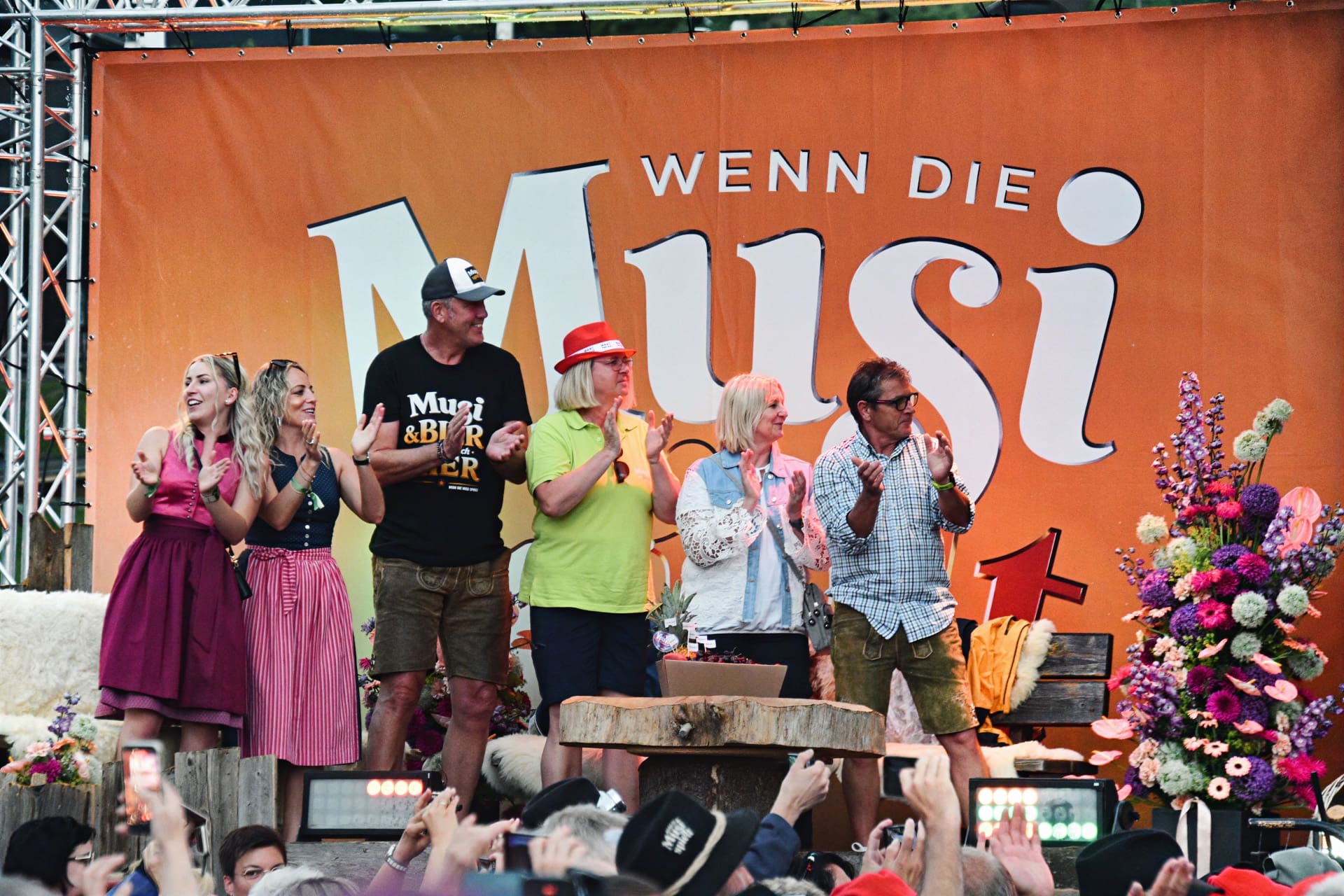 Mehrere Personen auf einer Bühne klatschen vor einem großen orangen Banner mit der Aufschrift Wenn die Musi