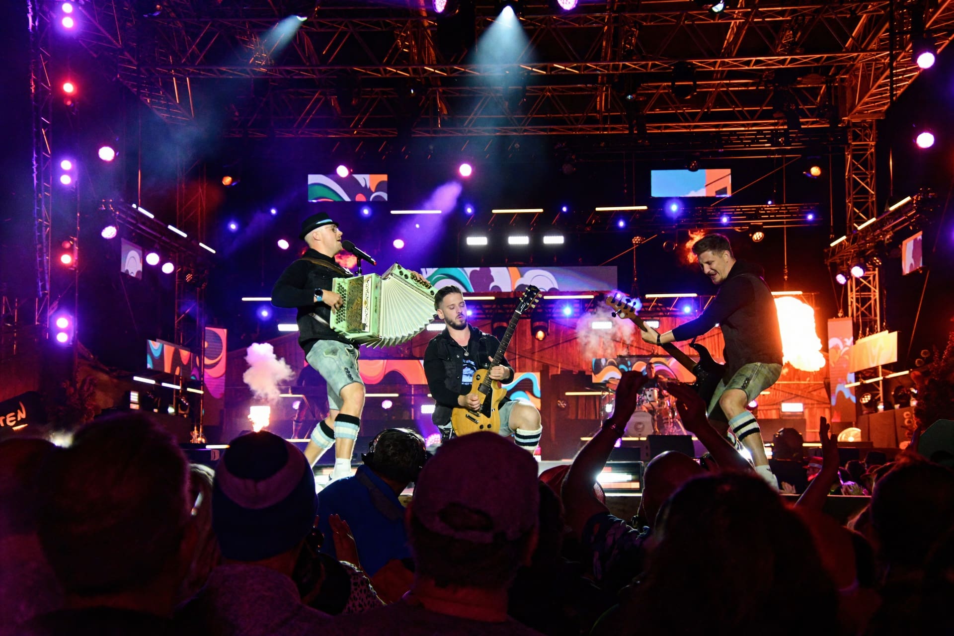 Drei Musiker mit Akkordeon und Gitarren auf einer beleuchteten Bühne, Publikum im Vordergrund.