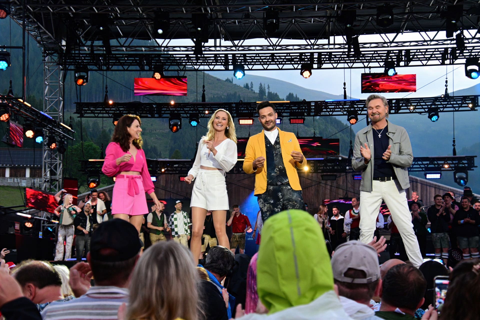 Vier Personen stehen applaudierend auf einer großen Bühne vor Publikum bei einem Open-Air-Konzert in den Bergen