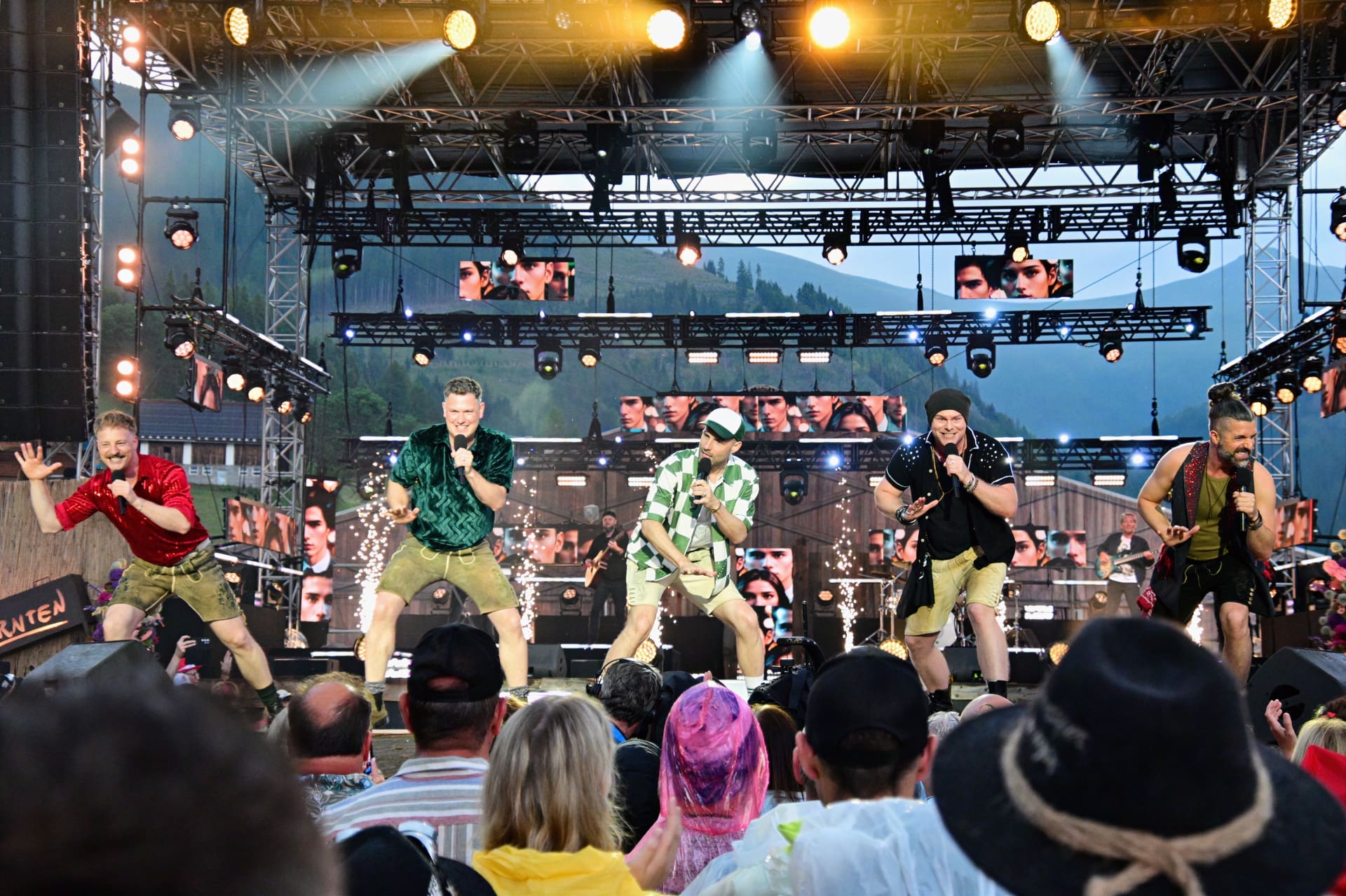 Vier männliche Performer singen und tanzen auf einer beleuchteten Open-Air-Bühne vor Publikum.