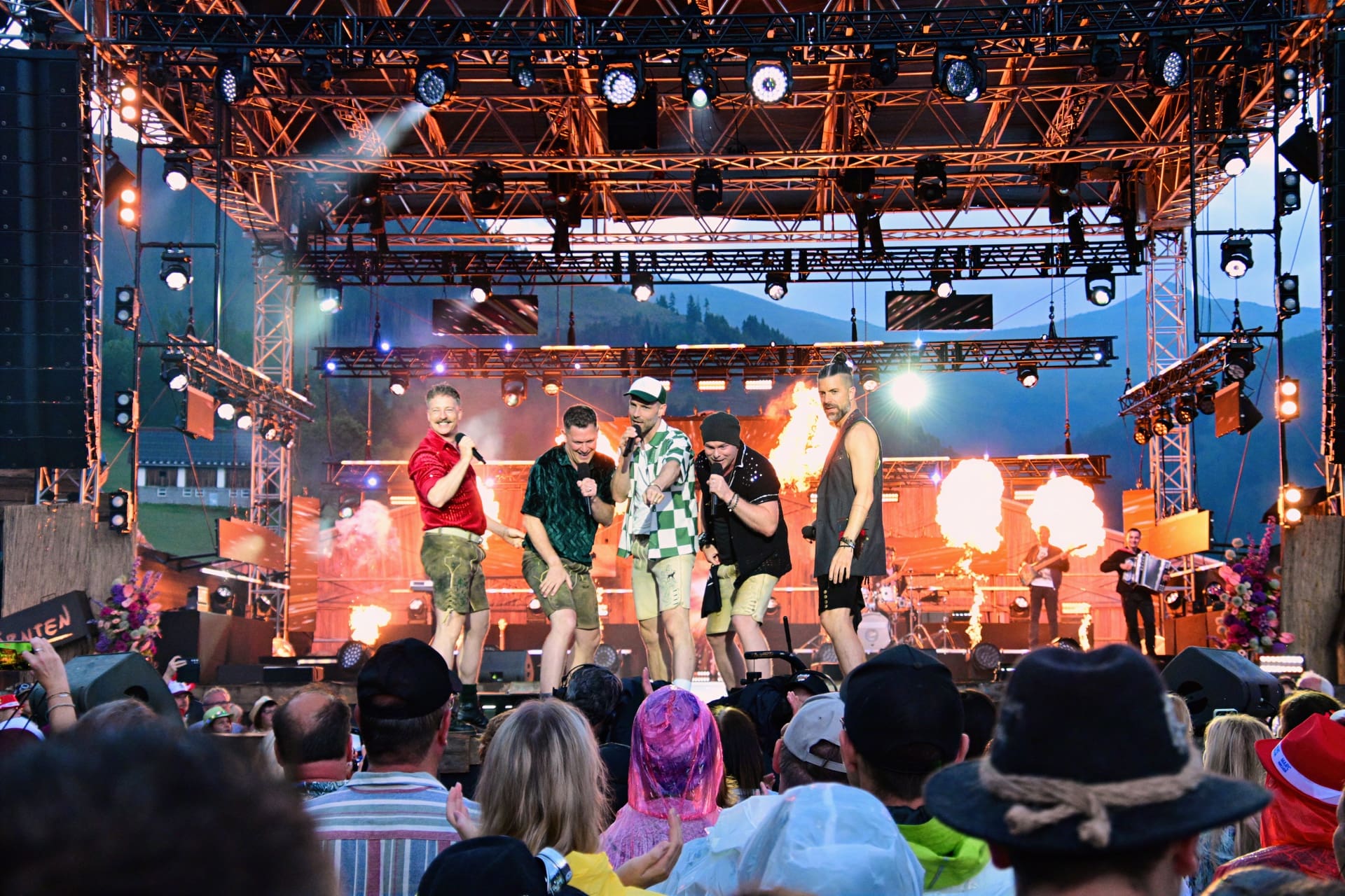 Mehrere Sänger in bunter Kleidung performen auf einer beleuchteten Bühne vor Publikum bei einem Open Air.