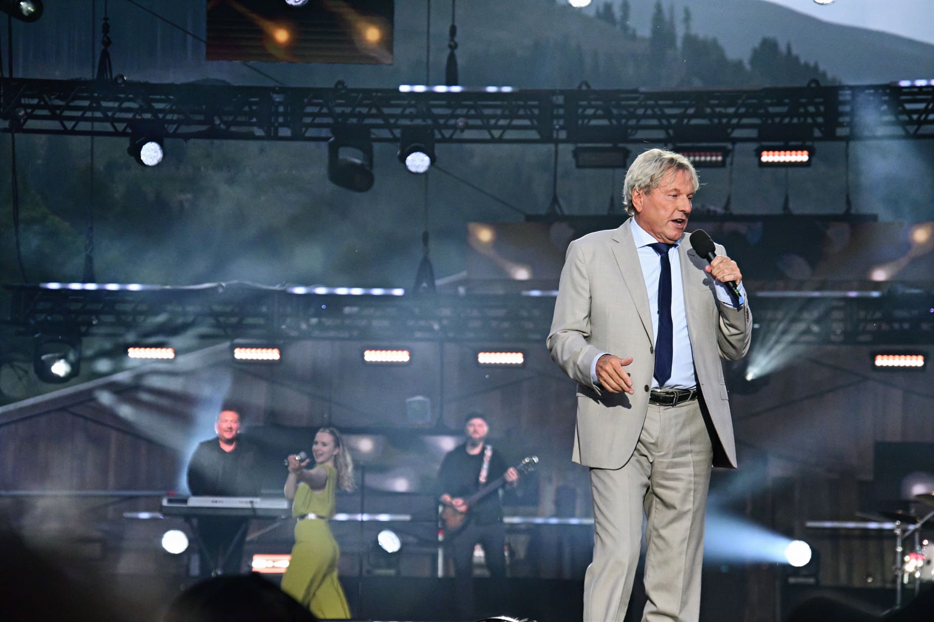 Sänger in hellem Anzug mit Mikrofon auf beleuchteter Bühne bei Open-Air-Konzert mit Band im Hintergrund