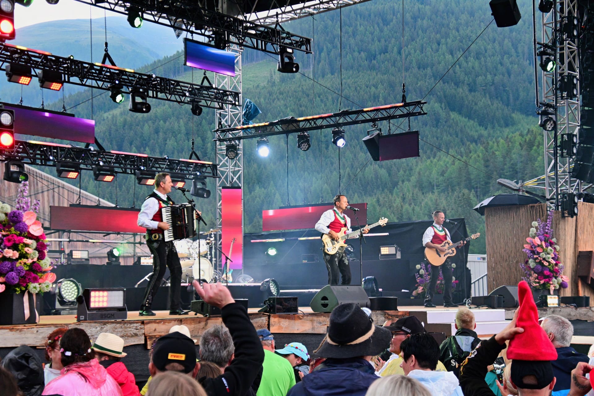 Drei Musiker mit Akkordeon und Gitarren auf einer Bühne vor Publikum und Bergen