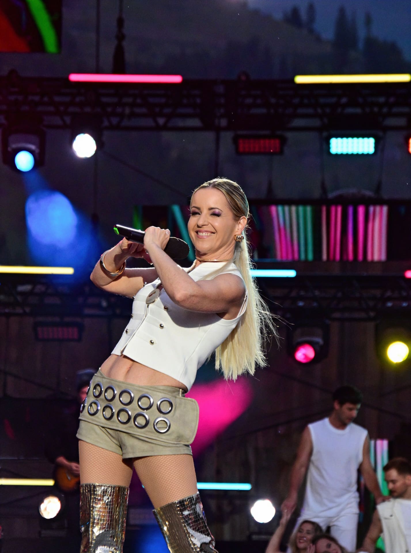 Blonde Sängerin in weißem Outfit singt mit Mikrofon auf einer beleuchteten Bühne.