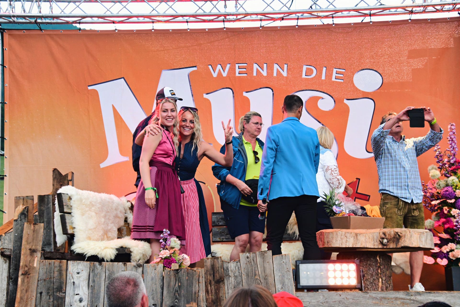 Zwei Frauen in Trachtenkleidern posieren lächelnd auf einer Bühne vor einem großen orangen Banner.