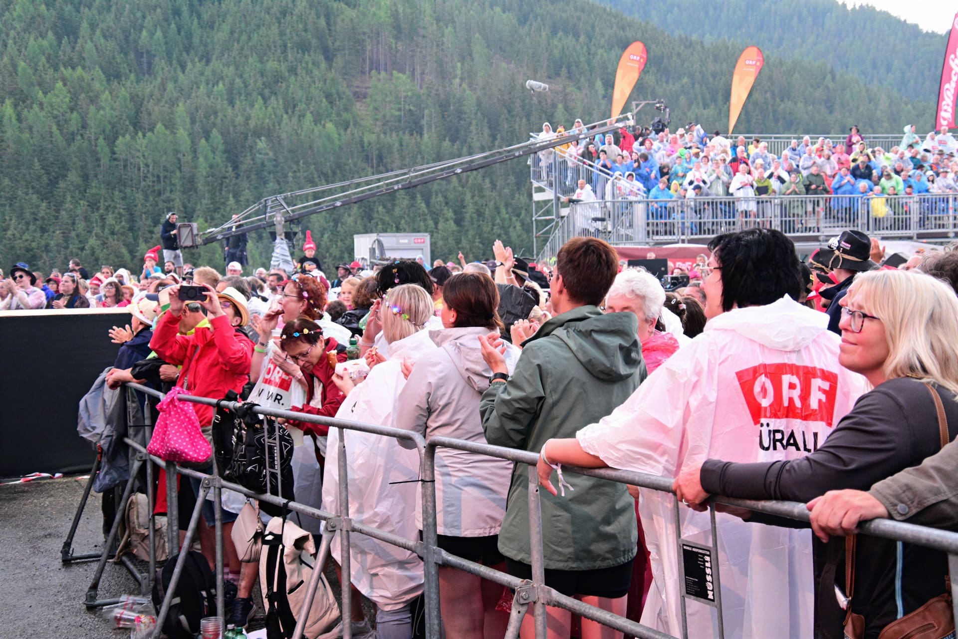 Zuschauer in Regenponchos stehen hinter Absperrungen beim Open-Air-Konzert in den Bergen