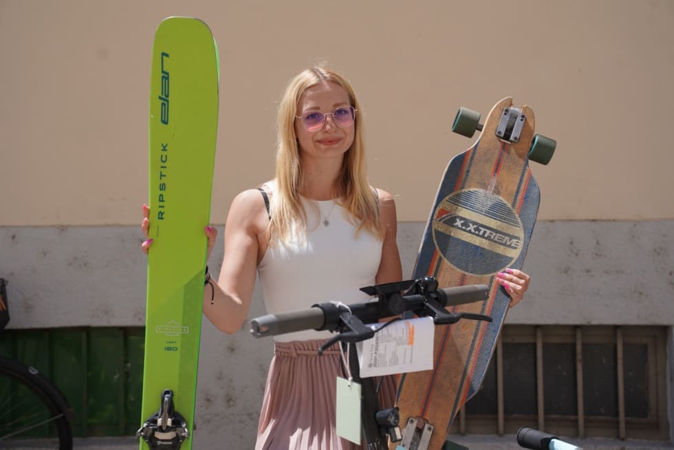 Eine blonde Frau hält einen grünen Ski und ein Longboard in den Händen.