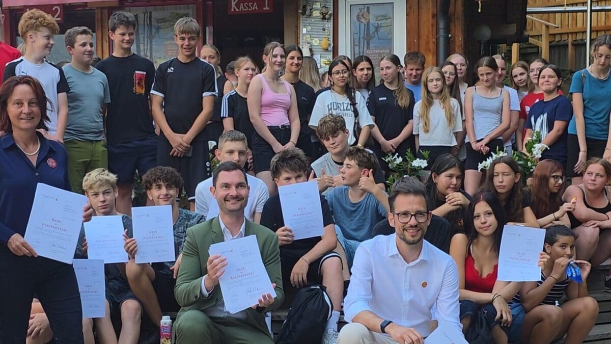 Gruppe von Schülerlotsinnen und Schülerlotsen mit Urkunden vor einem Holzhaus im Familywald Ossiach