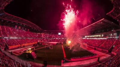 Nachtaufnahme eines voll besetzten Stadions mit rotem Licht und großem Feuerwerk über dem Spielfeld