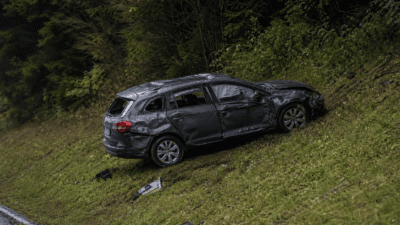 Schwer beschädigtes dunkles Auto liegt auf nasser grasbewachsener Böschung neben der Straße