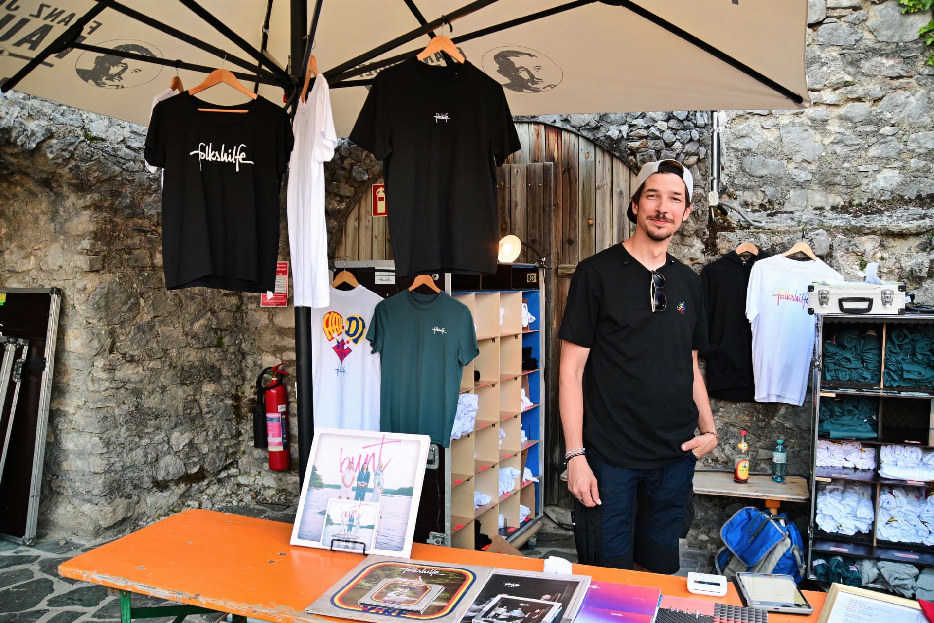 Mann steht an einem Verkaufsstand mit Folkshilfe T-Shirts und Schallplatten vor einer Steinmauer
