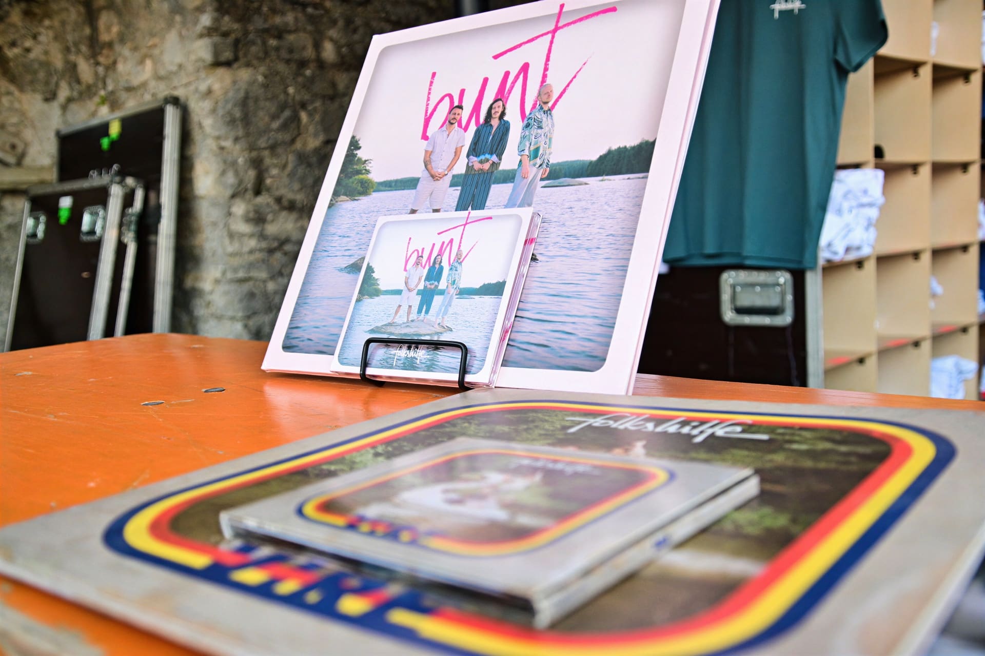 Großes pink gerahmtes Poster der Band Folkshilfe mit drei Männern am See und Schallplatten davor auf einem Tisch