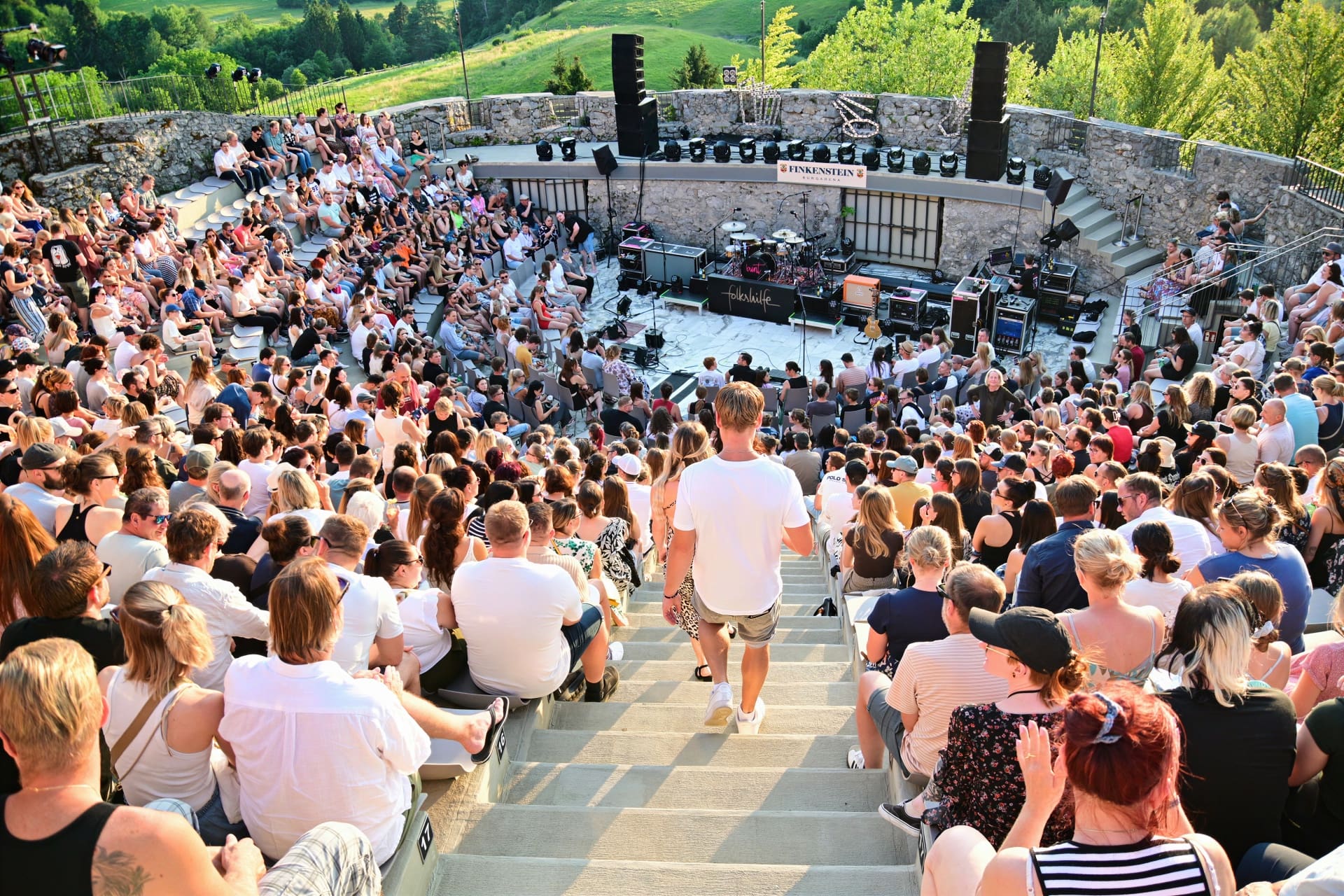 Große Menschenmenge sitzt auf Steinstufen einer Freiluftarena bei einem Konzert