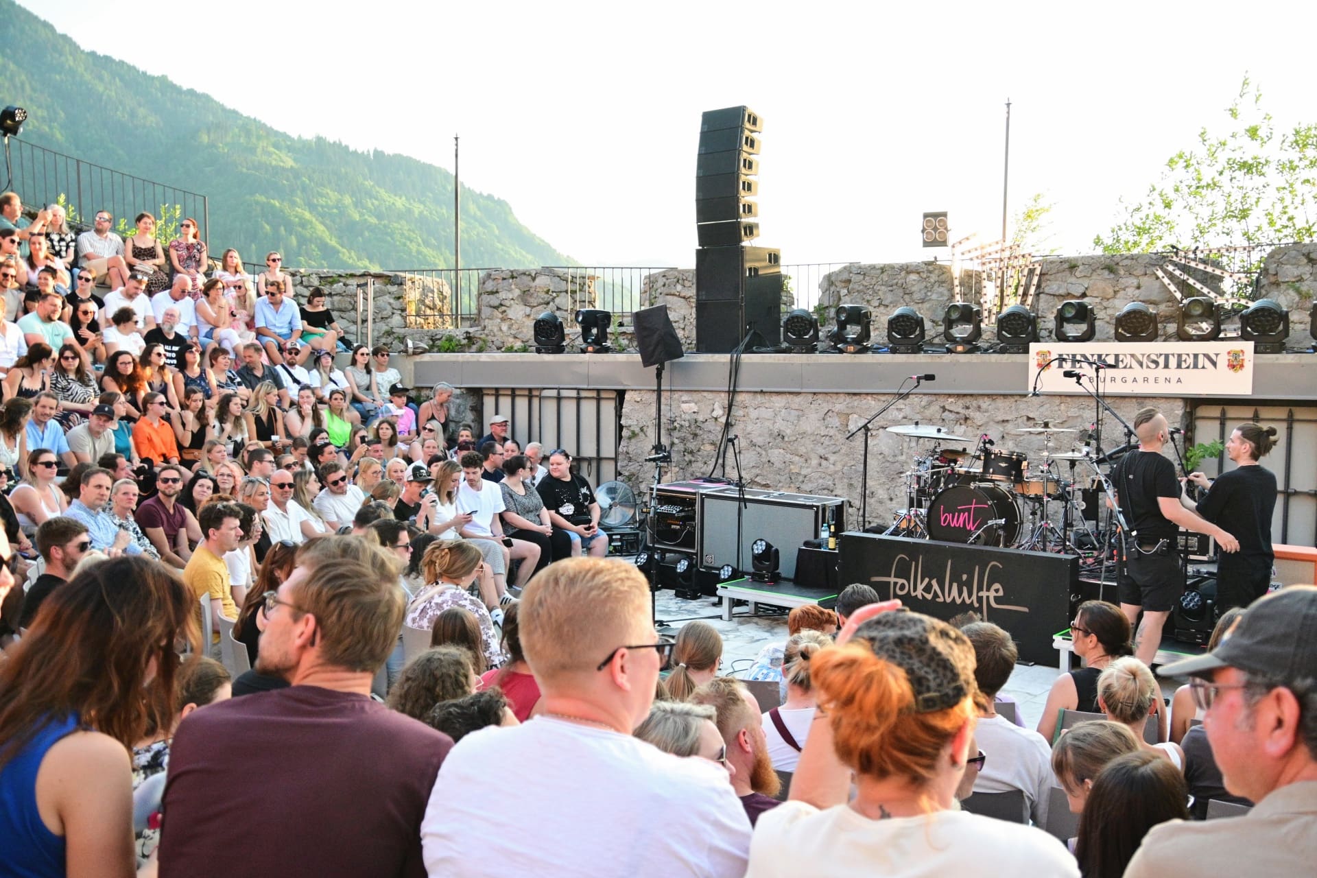Großes Publikum sitzt vor der Bühne mit Musikern und Lautsprechern in der steinernen Burgarena vor Bergen.