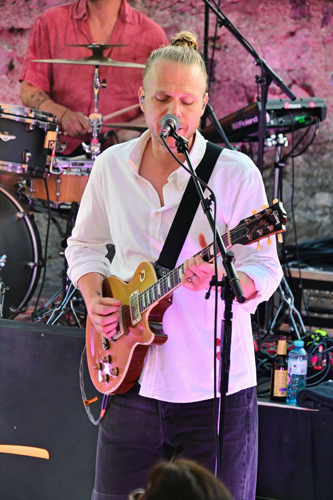 Blondhaariger Musiker mit Dutt spielt E-Gitarre und singt ins Mikrofon auf der Bühne.
