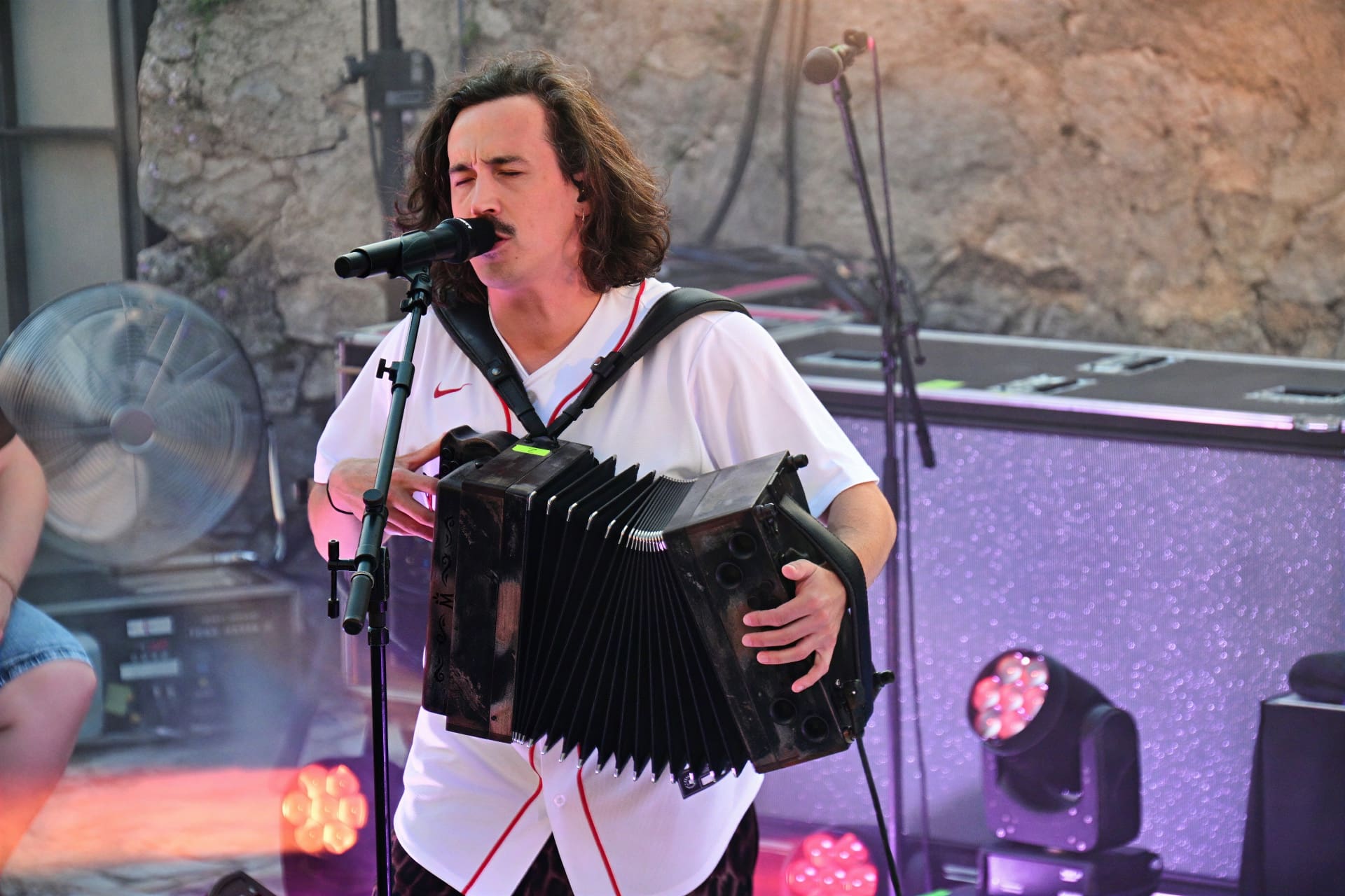 Ein Mann mit langen dunklen Haaren spielt Akkordeon und singt in ein Mikrofon auf der Bühne.