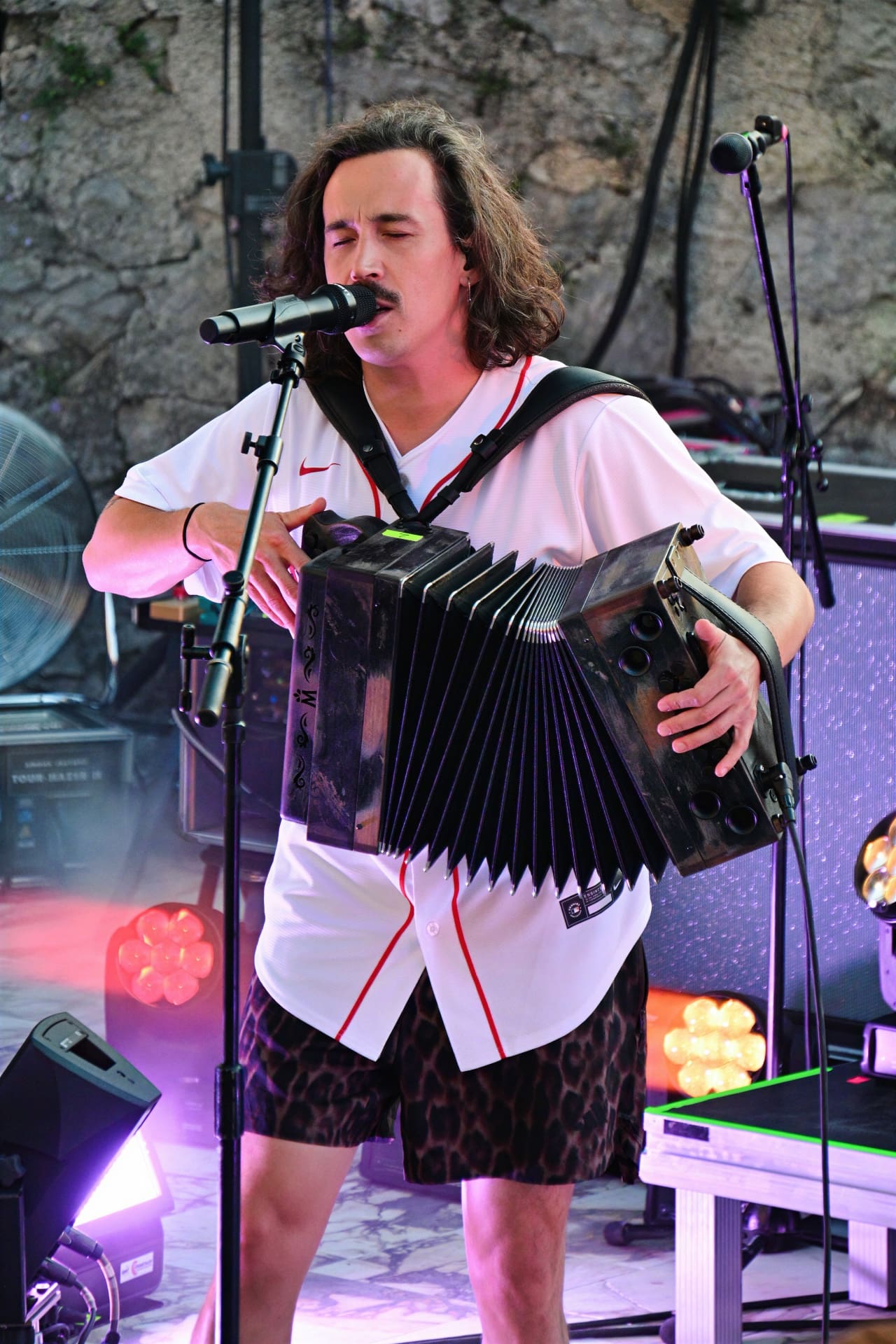 Ein Mann mit langen Haaren spielt Akkordeon und singt in ein Mikrofon auf einer Bühne.