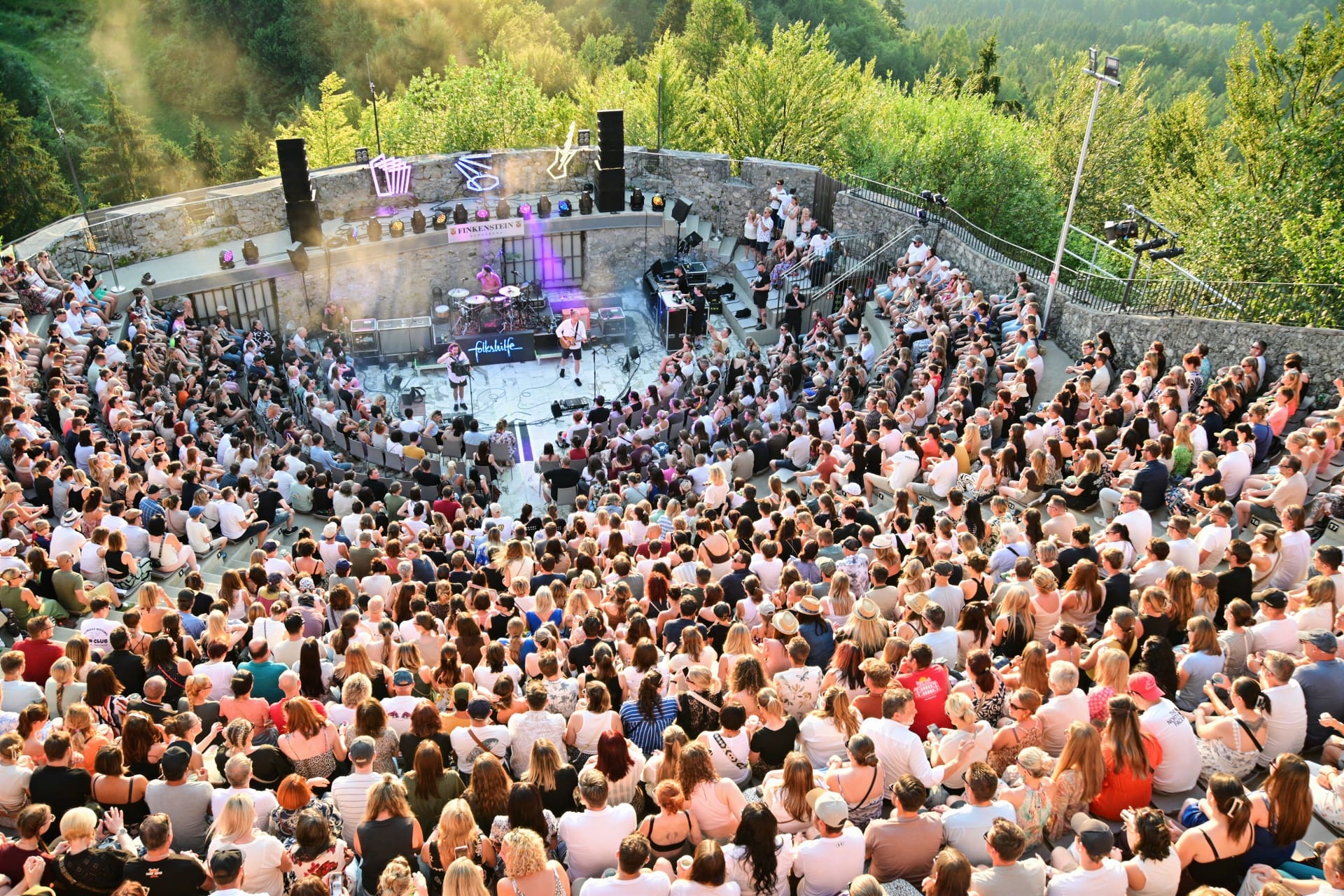 Große Menschenmenge sitzt in einer steinernen Freiluftarena bei einem Konzert von Folkshilfe.