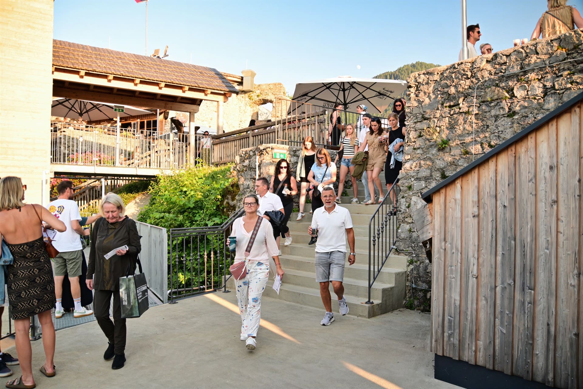 Menschen steigen Treppen an einer steinernen Burgarena mit Sonnenschirmen und Holzbauten hinauf und hinunter