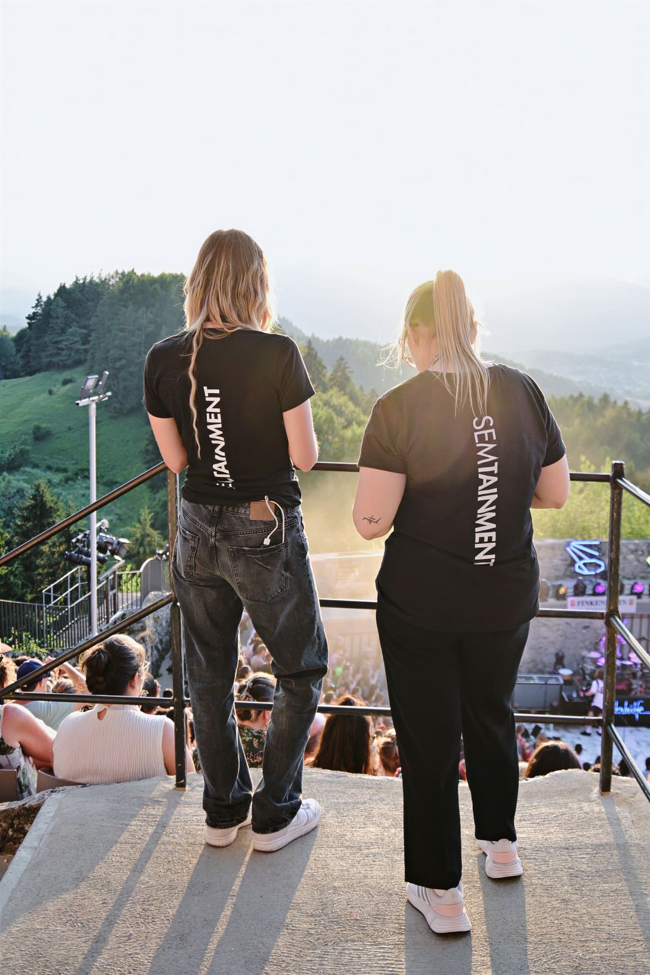 Zwei Frauen in schwarzen T-Shirts mit Semitainment-Aufdruck blicken auf ein Konzertpublikum in der Burgarena Finkenstein.