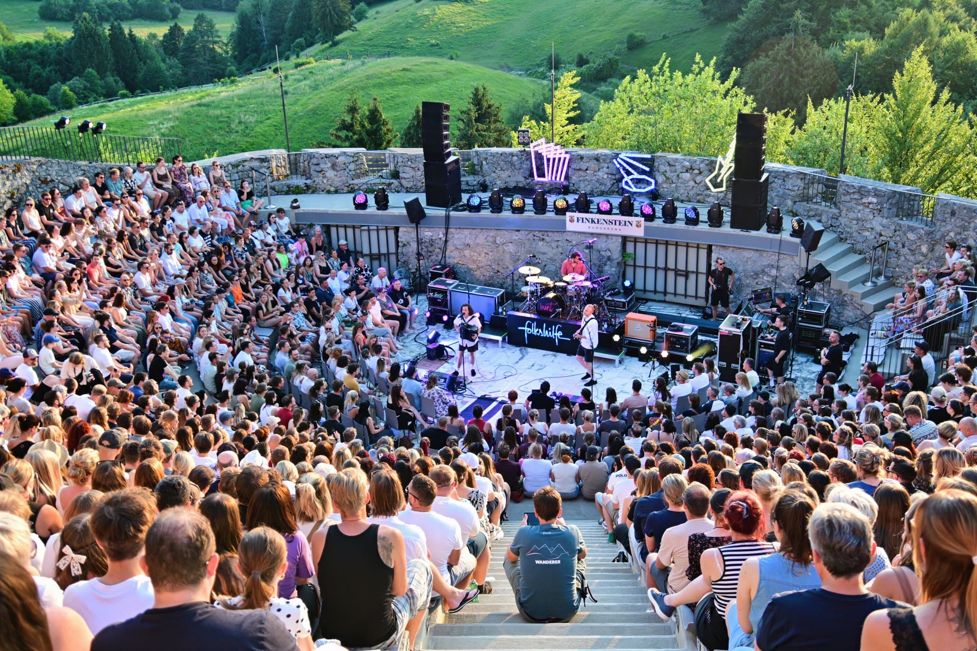 Große Menschenmenge sitzt auf Steinstufen vor einer Bühne bei einem Open-Air-Konzert in einer Burgarena.
