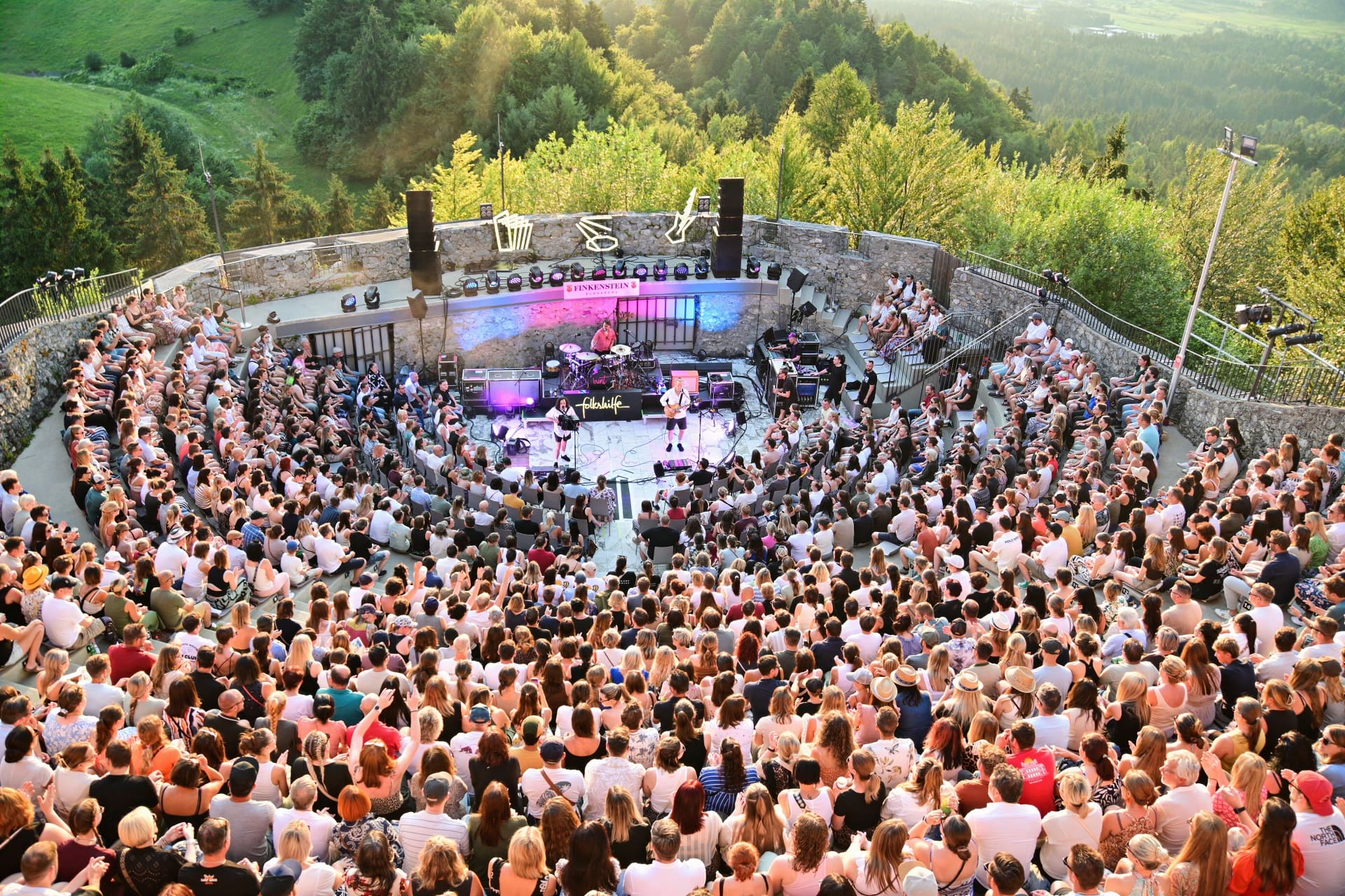 Große Menschenmenge sitzt auf Steinstufen vor einer Konzertbühne mit Band in bewaldeter Landschaft