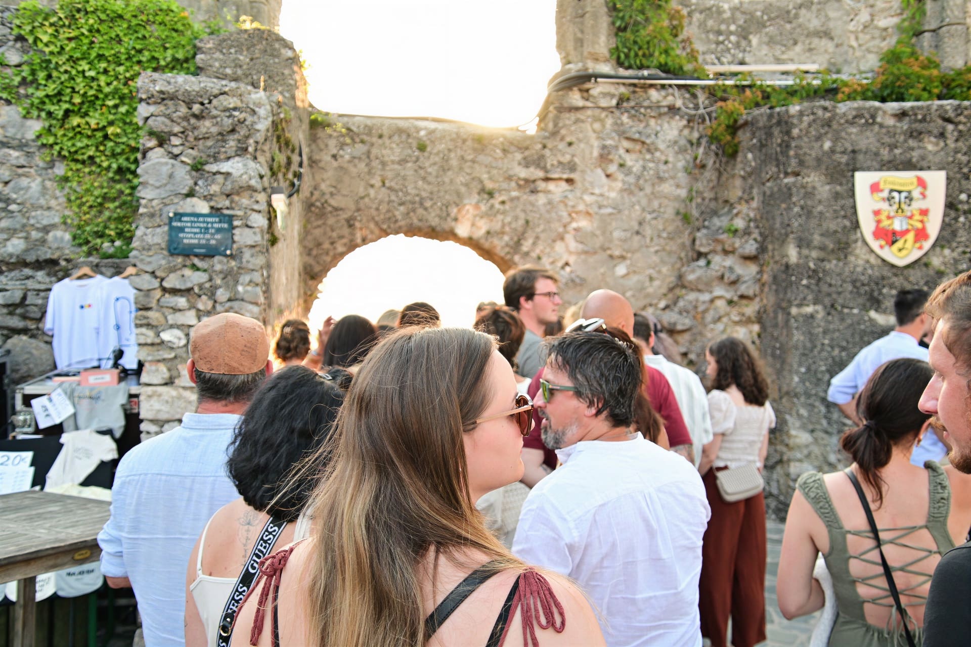 Menschenmenge vor alter Steinmauer mit Torbogen und Wappen bei Sonnenuntergang