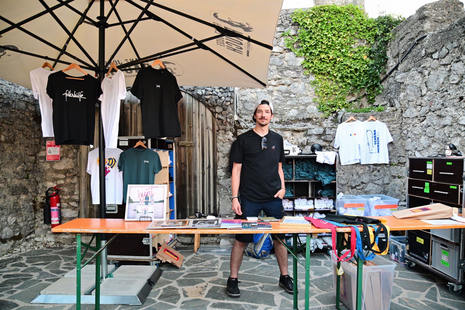 Ein Mann steht hinter einem Tisch mit T-Shirts und Schallplatten unter einem großen Sonnenschirm vor einer Steinmauer.