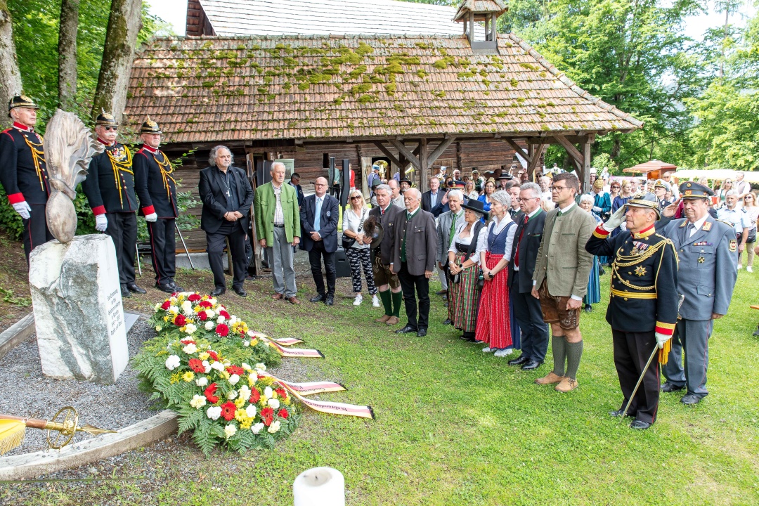 Menschen in traditioneller Tracht und Uniformen bei einer Kranzniederlegung vor einem Holzhaus im Freilichtmuseum Maria Saal