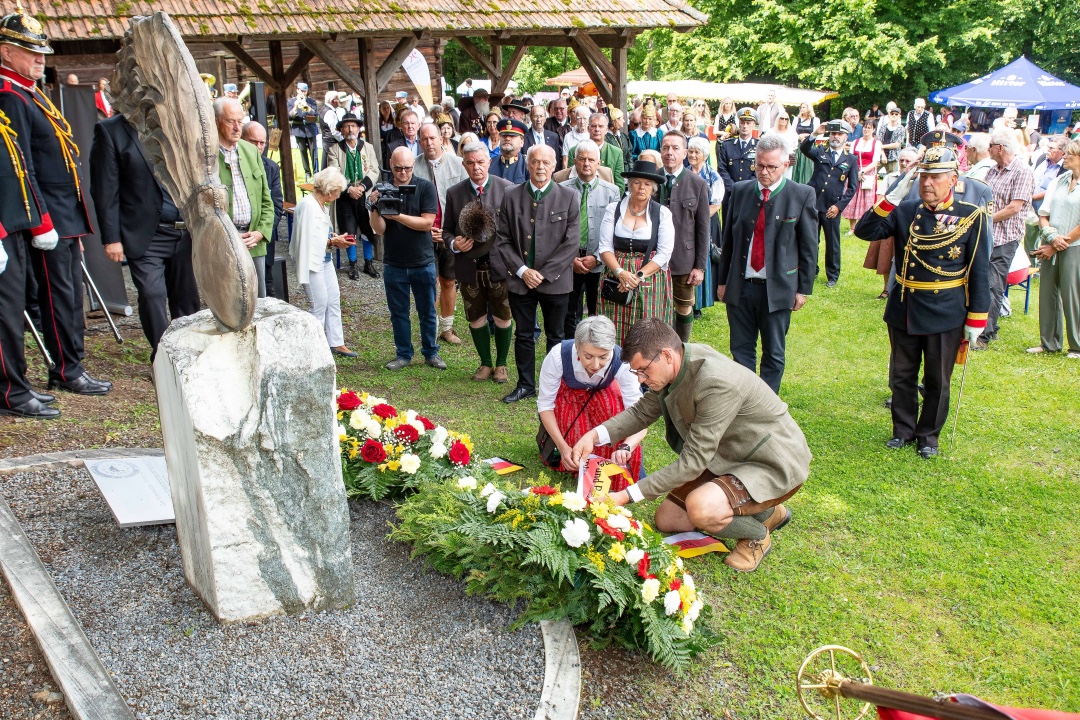 Mann in Tracht kniet vor Blumenkranz an Steinmonument im Freilichtmuseum