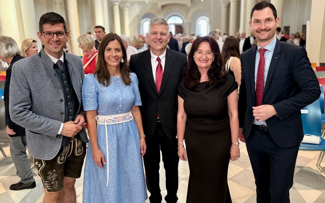 Fünf Personen in formeller und traditioneller Kleidung posieren in einem großen Saal mit Säulen.