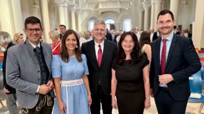 Fünf Personen in formeller und traditioneller Kleidung posieren in einem großen Saal mit Säulen.