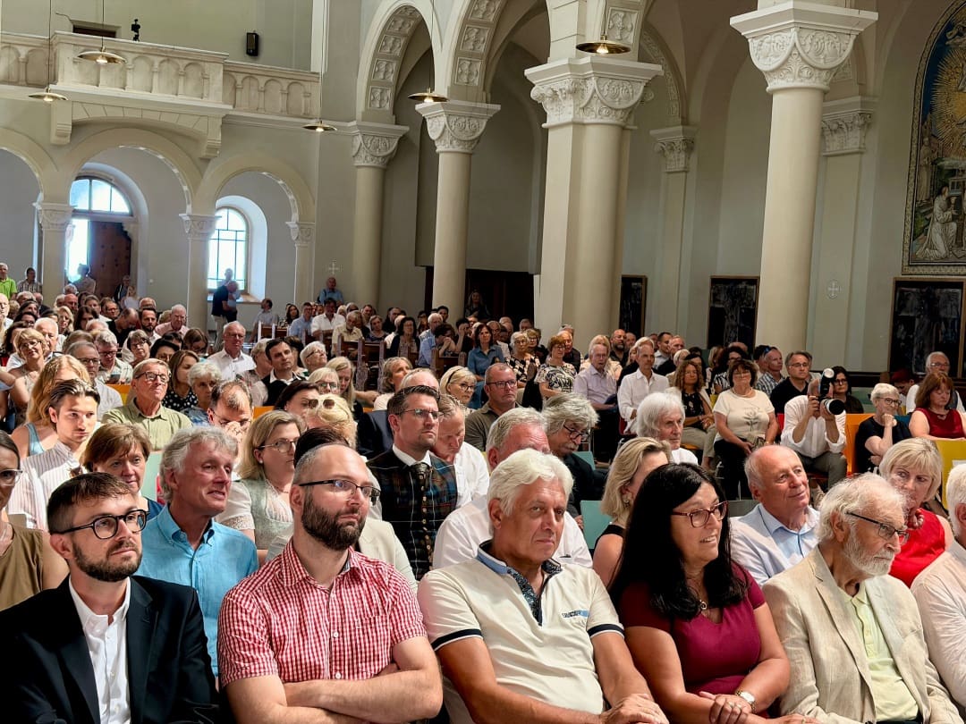 Zahlreiche Gäste sitzen in Reihen in einem prächtigen Kirchensaal mit Säulen und Bögen