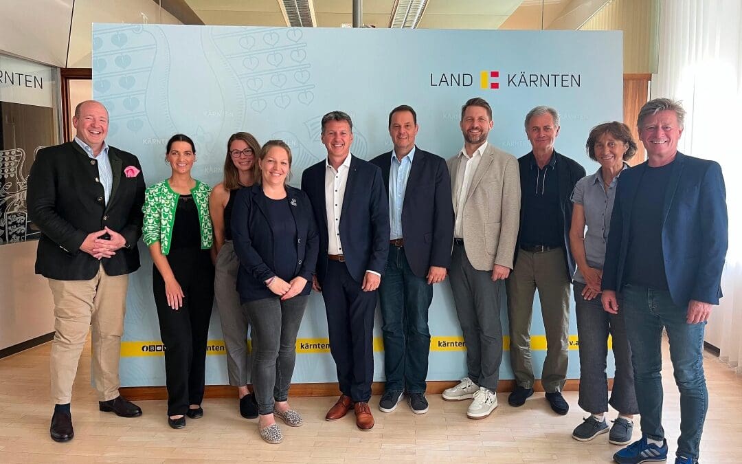 Neun Personen stehen lächelnd vor einem hellblauen Banner mit Logo des Landes Kärnten.