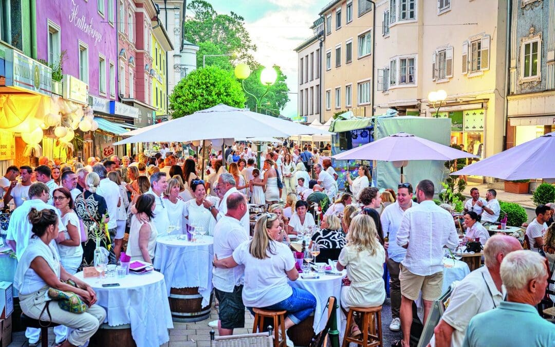 Menschen in weißer Kleidung sitzen an Tischen unter Sonnenschirmen in einer Fußgängerzone mit bunten Häusern.