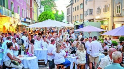 Menschen in weißer Kleidung sitzen an Tischen unter Sonnenschirmen in einer Fußgängerzone mit bunten Häusern.