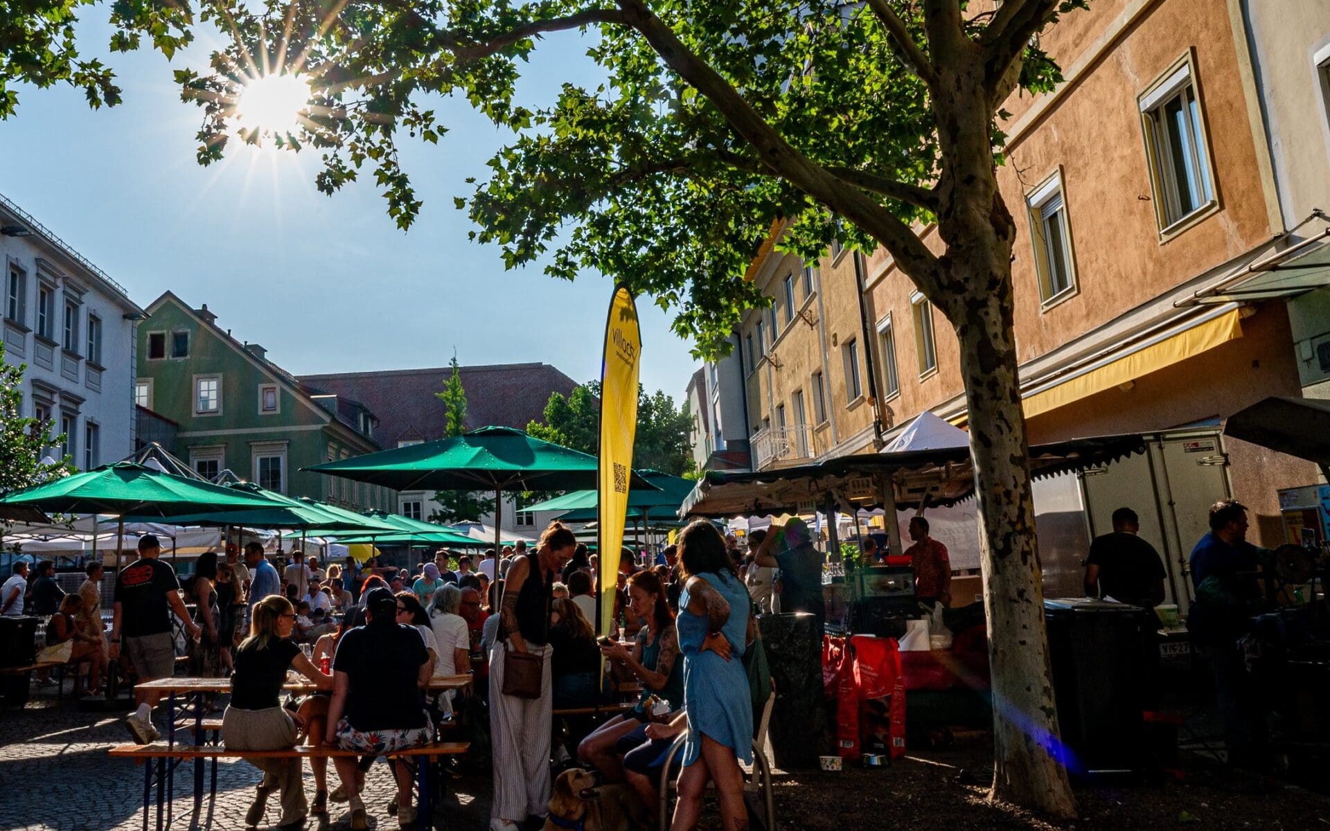 Menschen sitzen an Tischen unter grünen Sonnenschirmen auf einem belebten Streetfood-Markt an einem sonnigen Tag