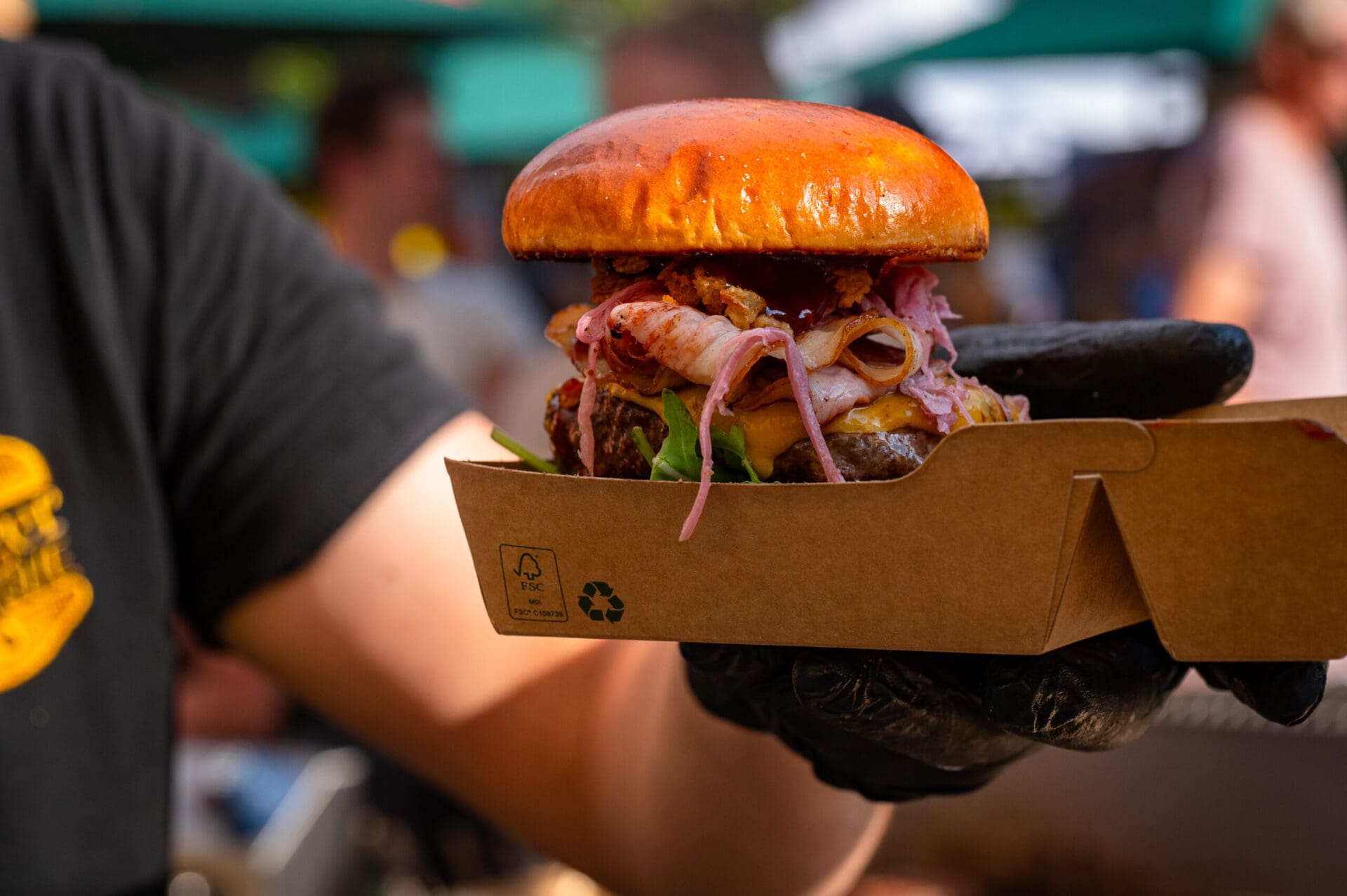 Eine Person mit schwarzen Handschuhen hält einen Burger mit Bacon und Käse in einer Pappbox.