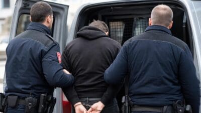 Zwei Polizisten in Uniform geleiten einen mit Handschellen gefesselten Mann in einen Polizeiwagen