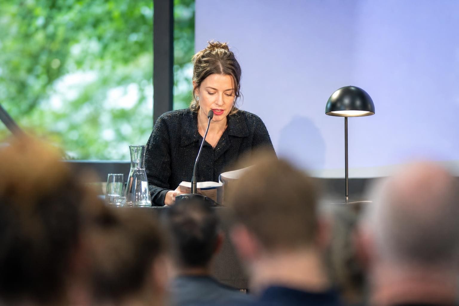 Eine Frau liest an einem Tisch mit Mikrofon und Lampe vor Publikum
