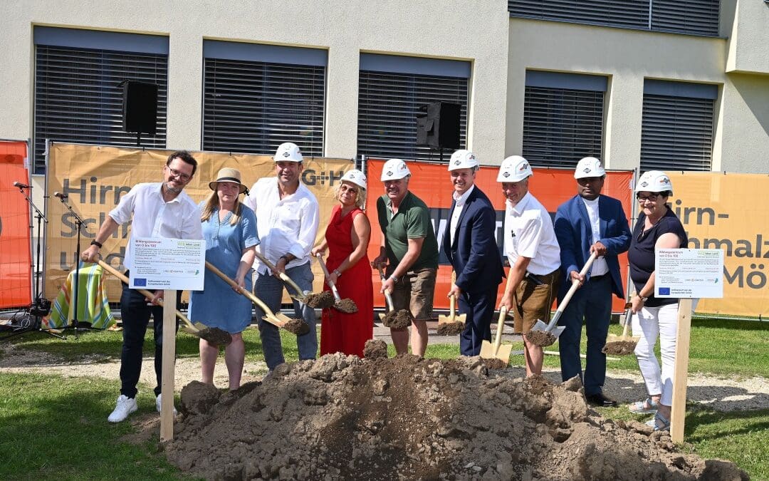Gruppe von Menschen mit Schutzhelmen und Schaufeln beim Spatenstich vor einem Gebäude