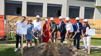 Gruppe von Menschen mit Schutzhelmen und Schaufeln beim Spatenstich vor einem Gebäude