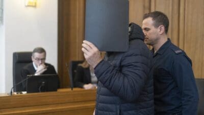 Angeklagter verdeckt sein Gesicht mit einem Ordner neben Polizist im Gerichtssaal