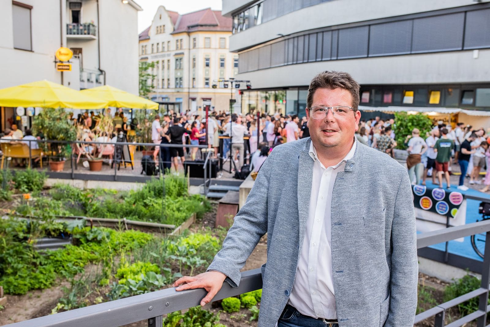 Ein Mann in grauem Sakko lehnt an einem Geländer vor einer Menschenansammlung mit gelben Sonnenschirmen.