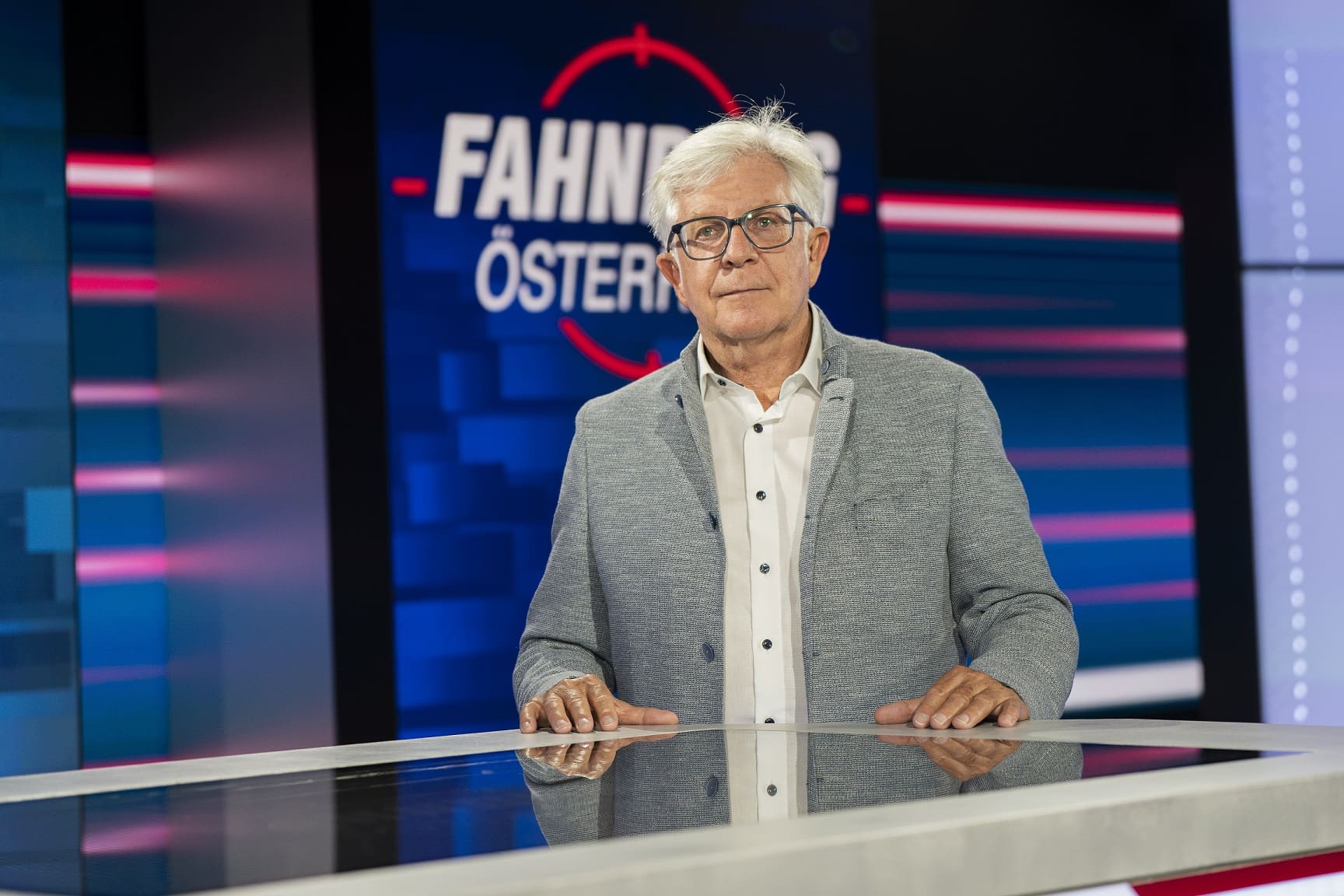 Älterer Mann mit weißem Haar und Brille steht hinter einem Tisch im Fernsehstudio