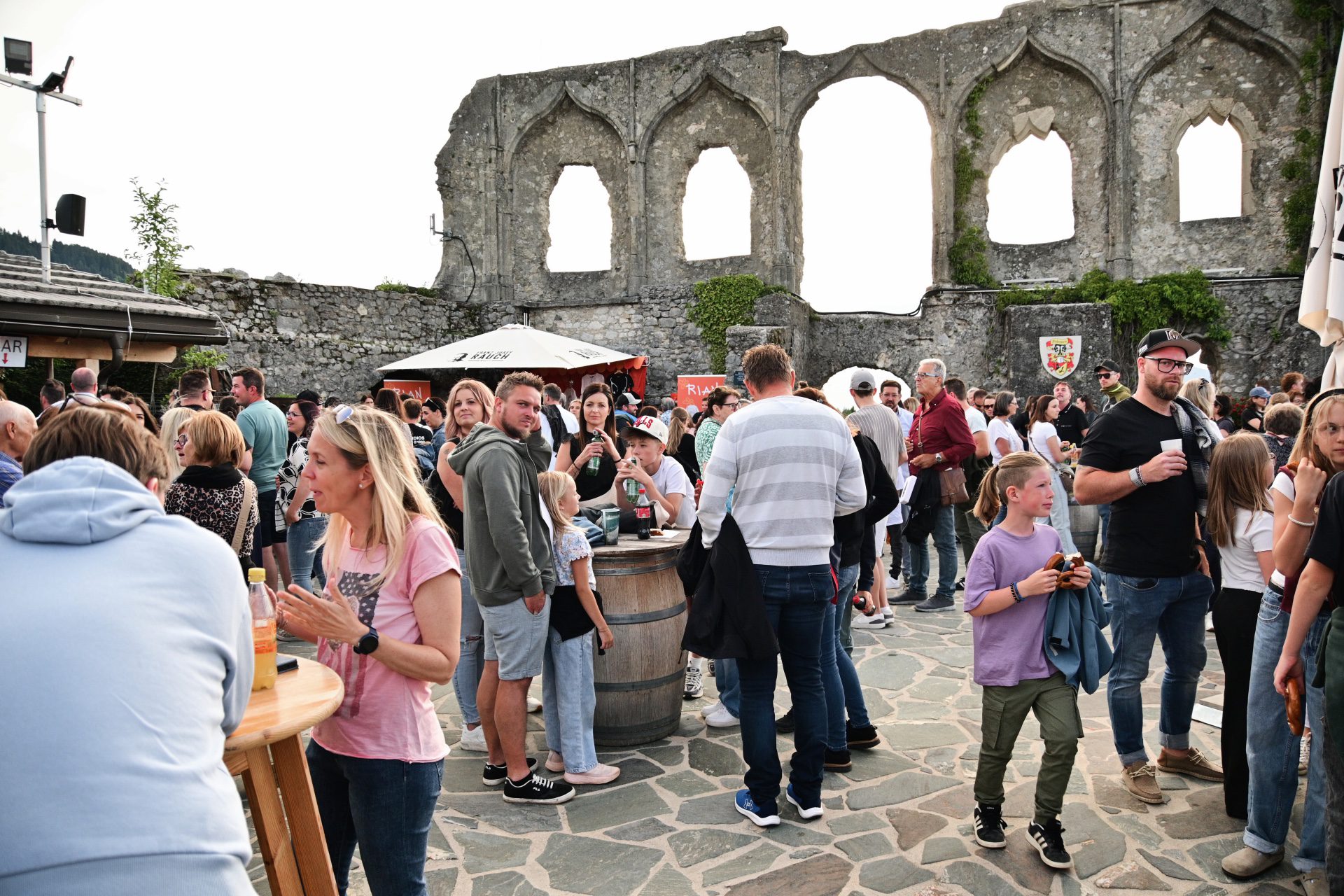 Menschenmenge bei einem Fest vor historischen Steinruinen mit Holzfass als Tisch
